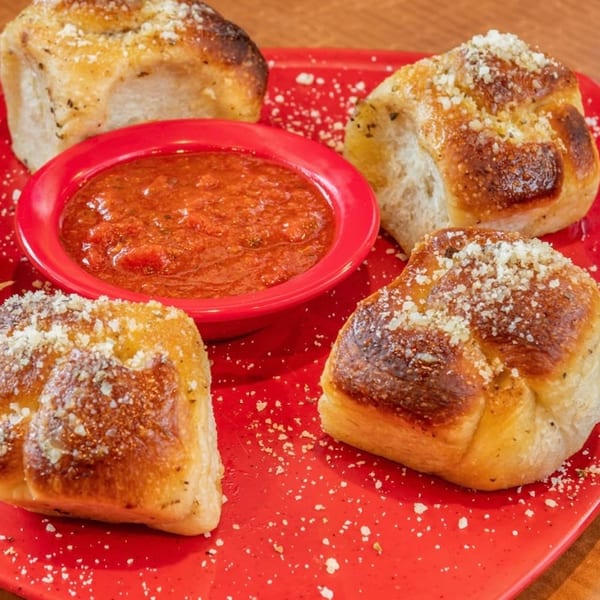 Four golden-brown garlic knots with parmesan cheese and a red bowl of marinara sauce on a red plate.